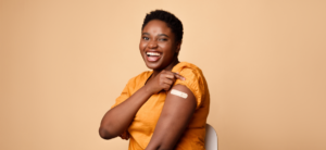 Black woman in a yellow shirt, rolling up her sleeve to show off her bandage from her covid-19 vaccine. She's smiling and standing in front of a tan background