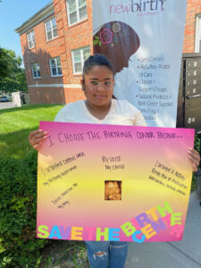 Carla Moralez holding a pink and yellow poster board sign that reads "I choose the Birthing Center because... I wanted control over my birthing experience, my voice my choice, I wanted a natural birth free of medication and western medical practices, better transition for my baby." In bright bold letters at the bottom of the sign, it reads "Save the Birth Center" She is standing in front of an informational sign about the New Birth Company